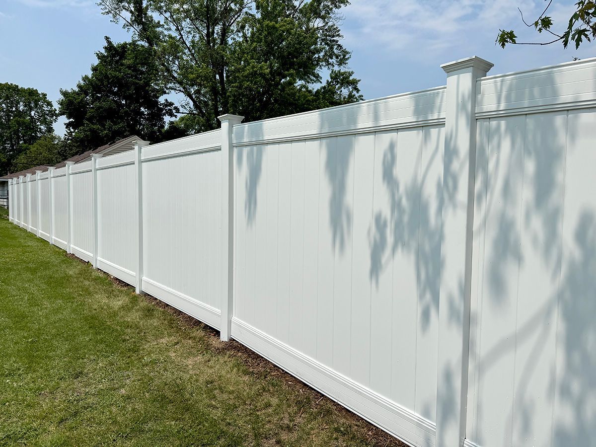 A long, white, solid vinyl privacy fence stands in a grassy lawn under a clear blue sky with dappled sunlight.