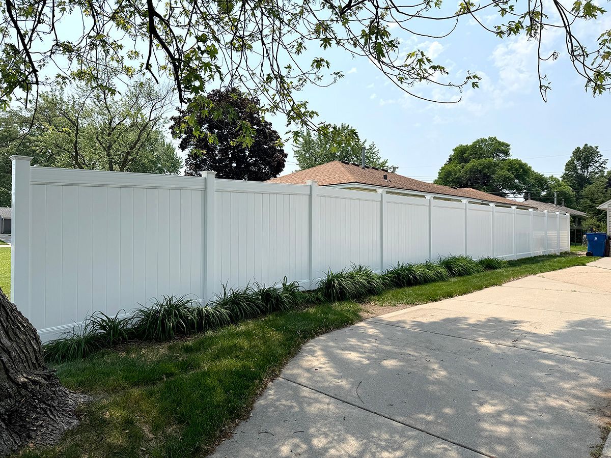 A long, white vinyl privacy fence runs alongside a concrete driveway on a sunny day with green landscaping at the base.