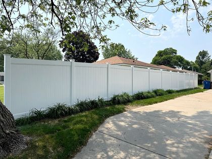 A long, solid white vinyl privacy fence lines a paved driveway on a sunny day with green grass and trees in the background.