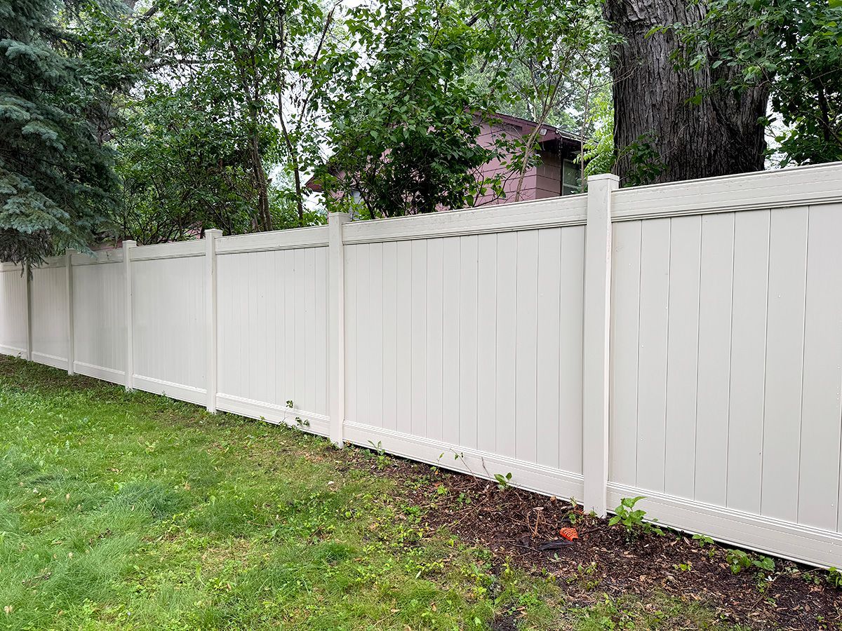 A tall, solid tan vinyl privacy fence borders a grassy backyard with trees and a house visible in the background.