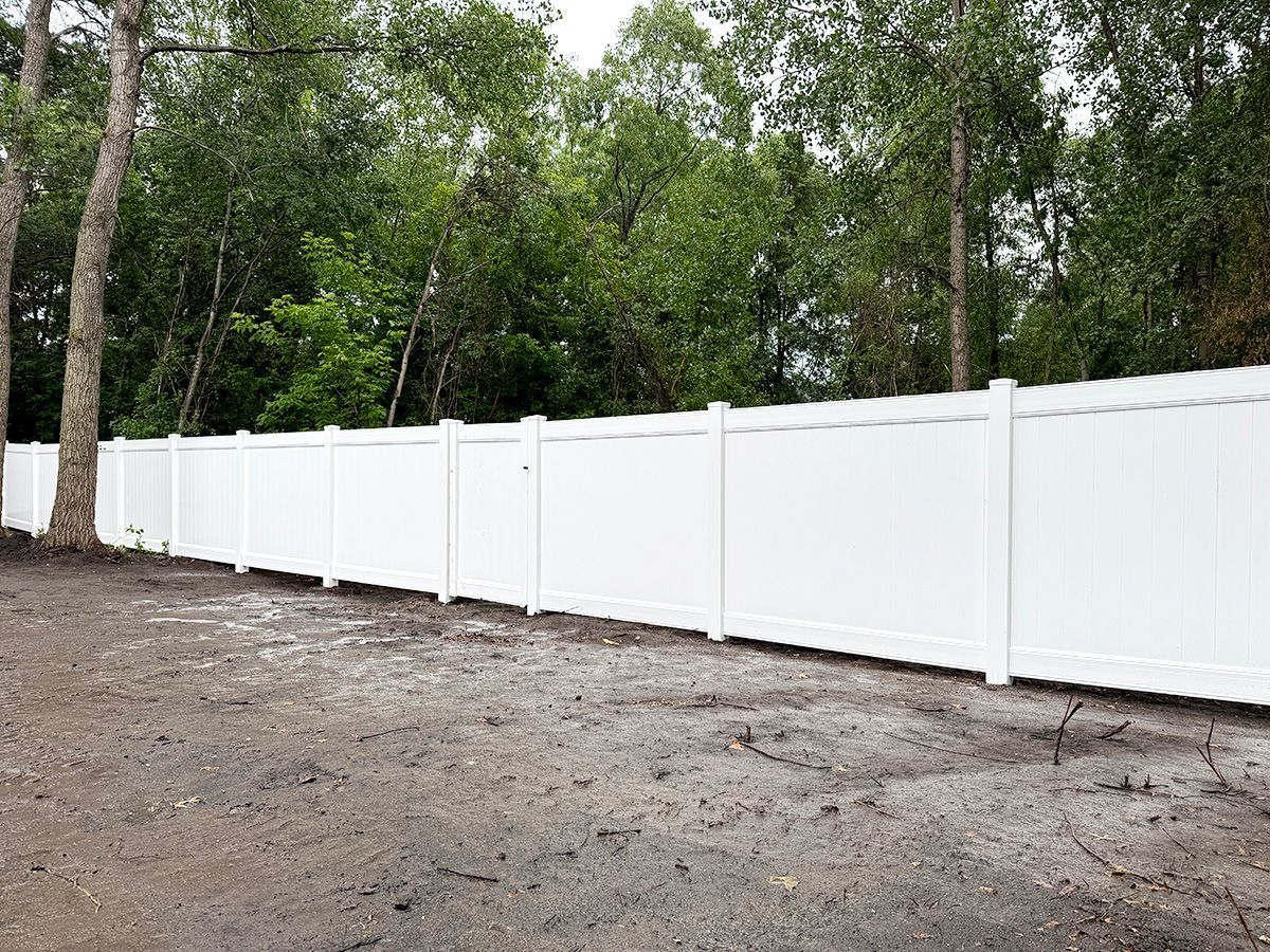 A long, white vinyl privacy fence stands in front of a dense forest with a dirt ground in the foreground.