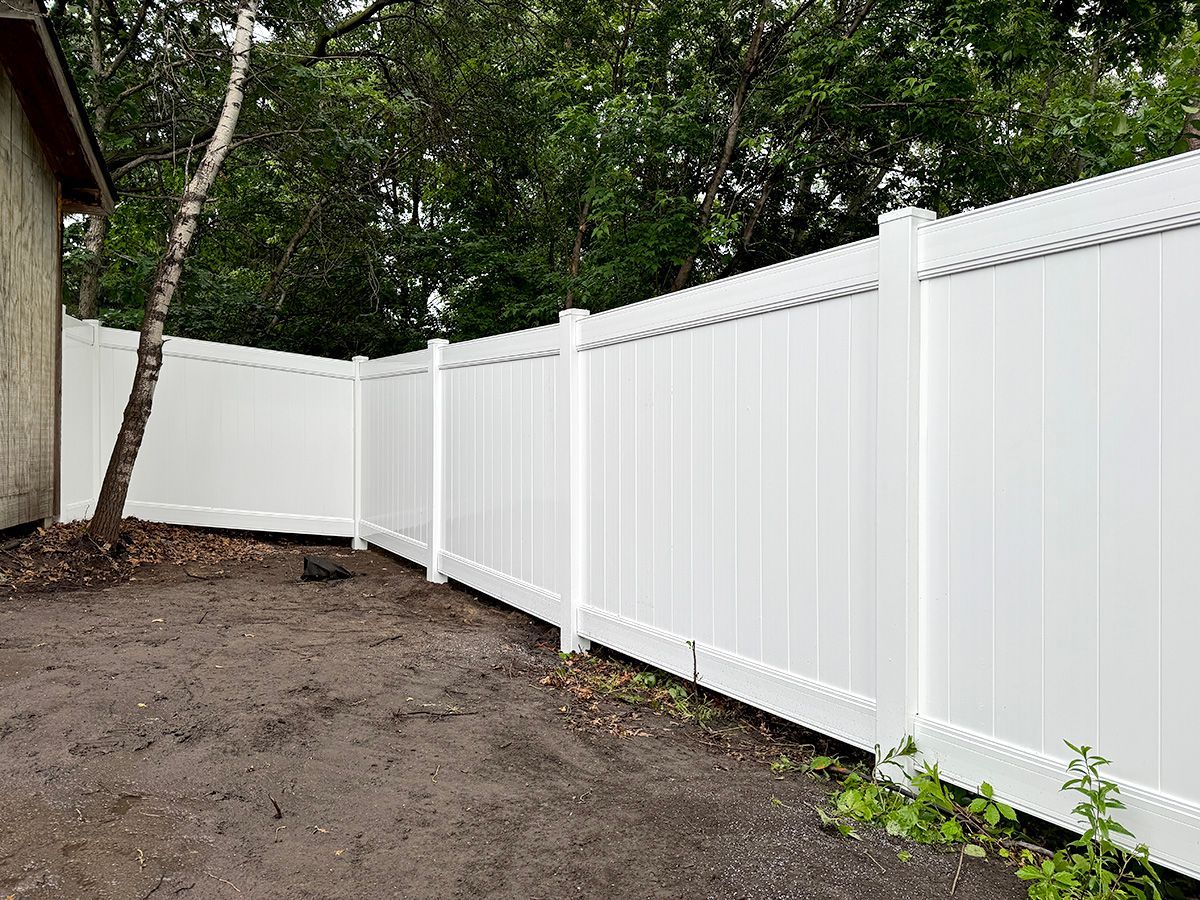A white vinyl privacy fence installed in a backyard with dirt ground and trees in the background.