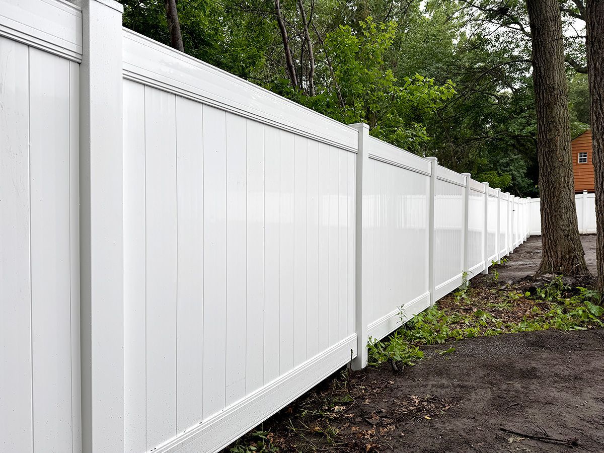 A long, white vinyl privacy fence lines the edge of a wooded area with dirt ground.