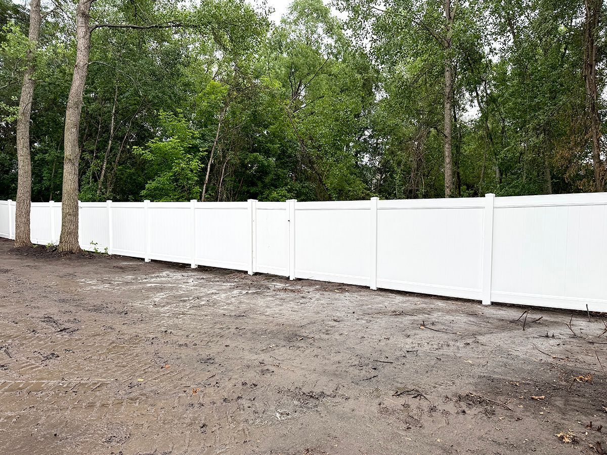 A white vinyl privacy fence stands in a dirt yard in front of a line of trees.