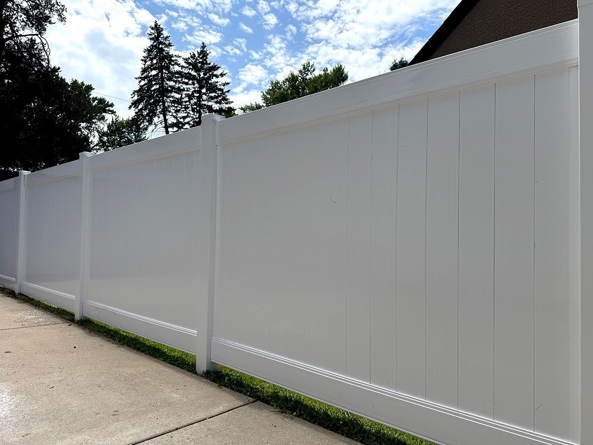 A white vinyl privacy fence lines a concrete path with trees and a blue sky in the background.