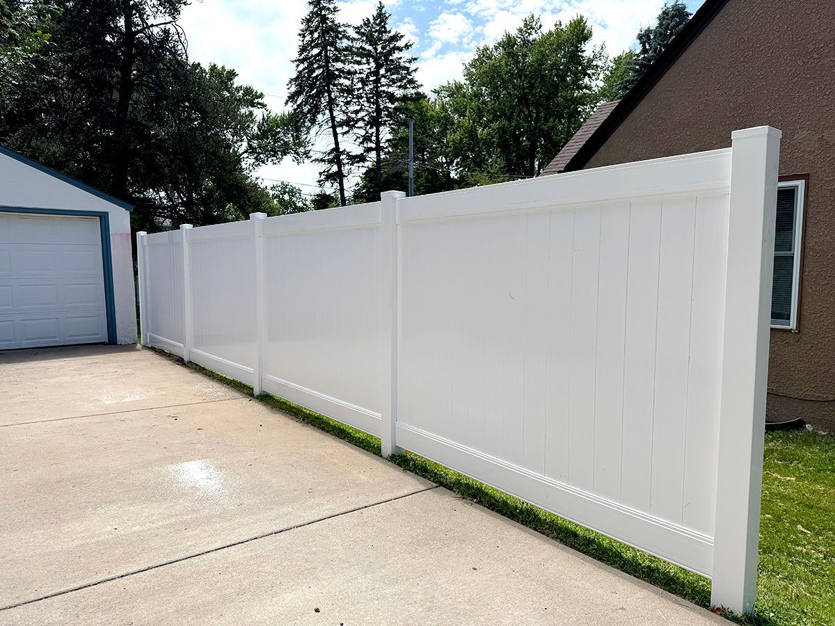 A long, solid white vinyl privacy fence stands between a concrete driveway and a grassy lawn on a sunny day.