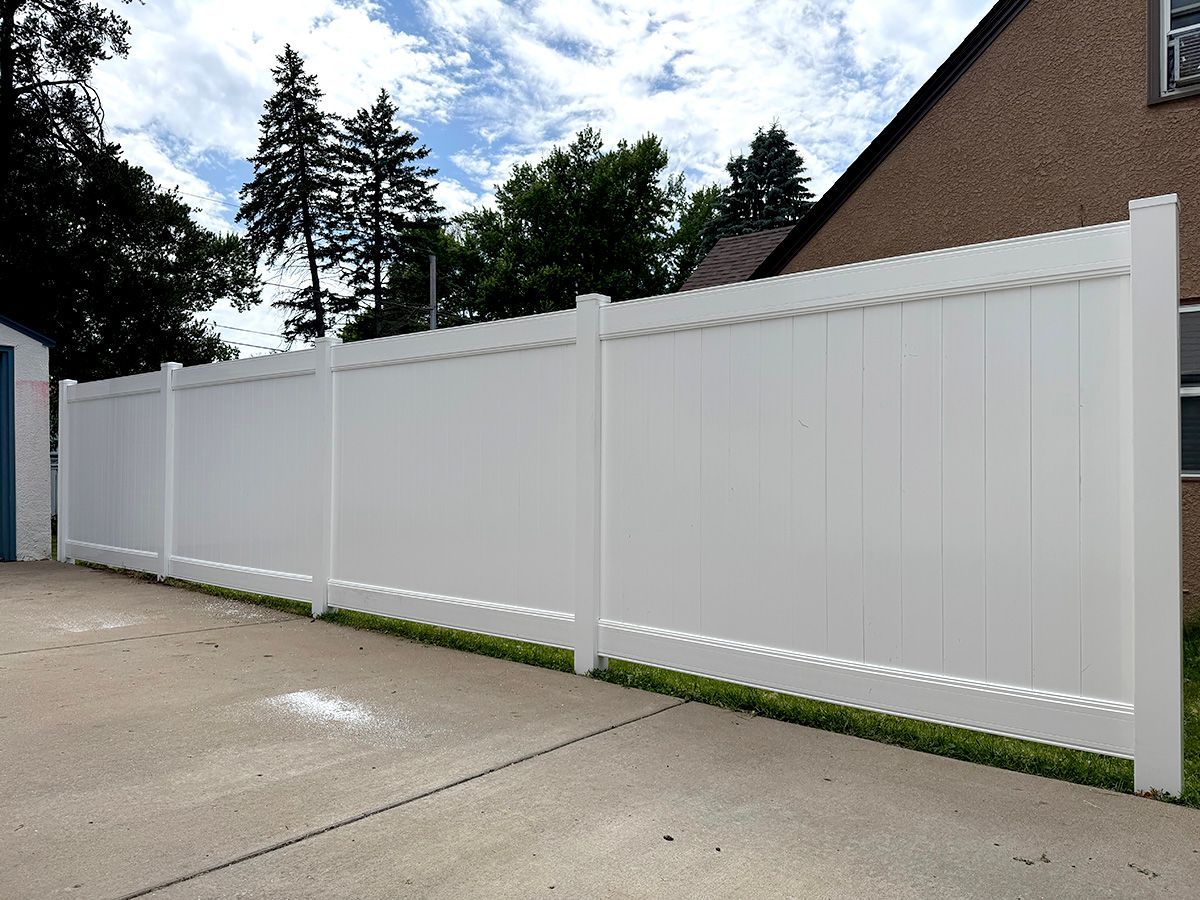 A tall white vinyl privacy fence lines a concrete driveway next to a brick house under a partly cloudy sky.