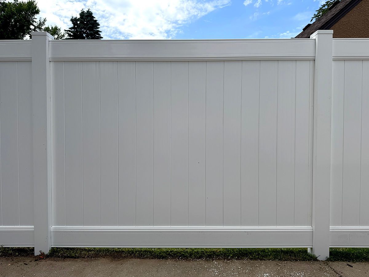 A white, solid vinyl privacy fence stands outdoors on a paved surface against a blue sky with clouds.