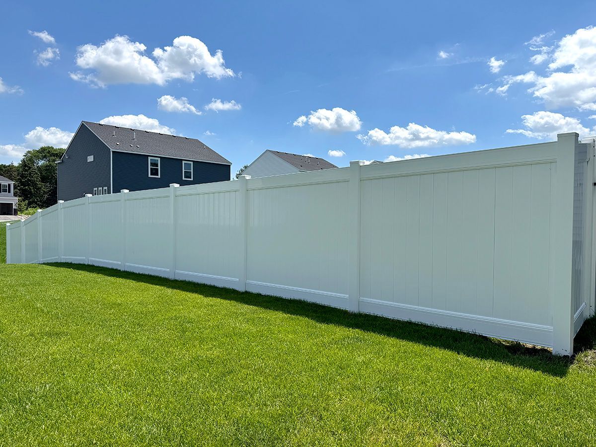 A long, solid white vinyl privacy fence stands in a grassy yard under a bright blue sky with scattered clouds.
