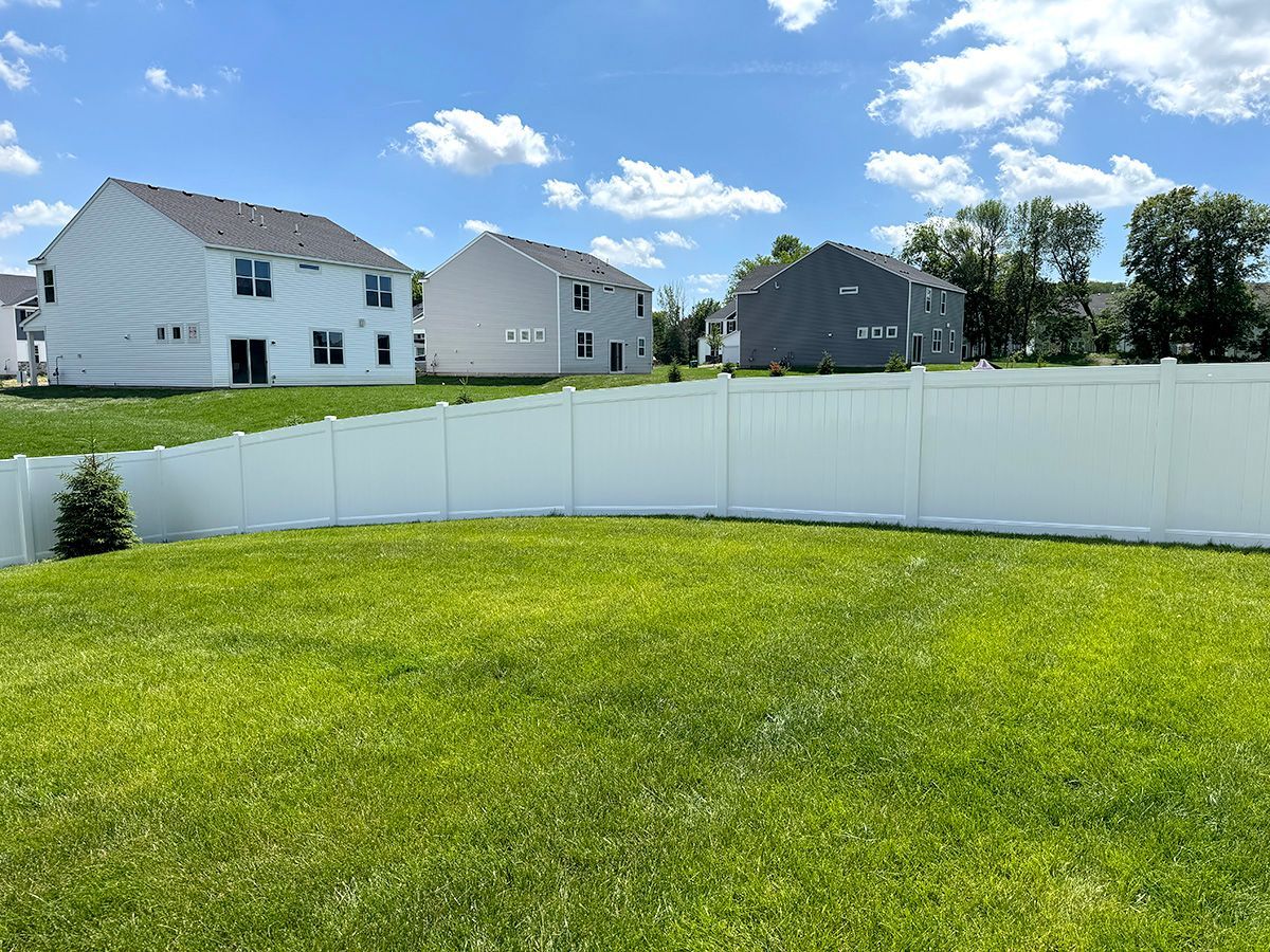 A lush green backyard featuring a white privacy fence, with several suburban houses in the background under a blue sky.