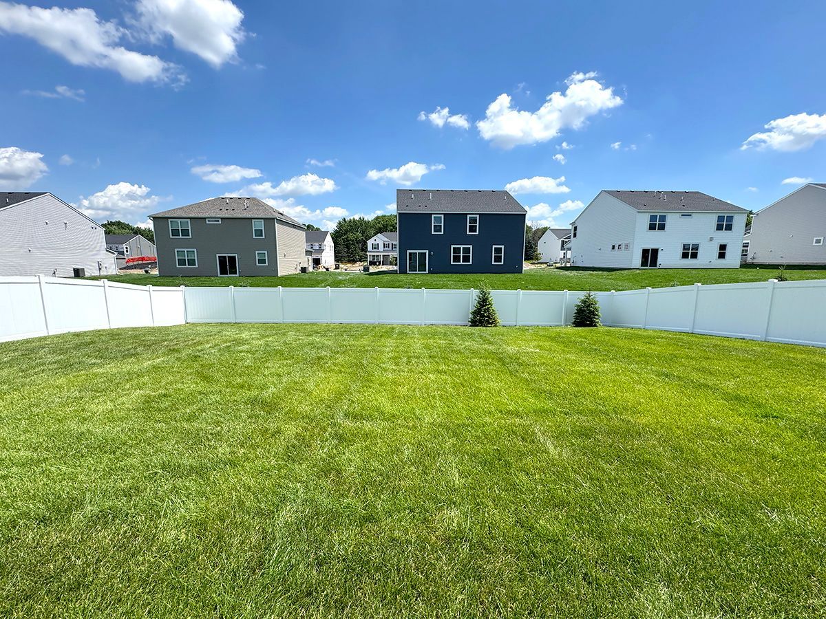A grassy backyard enclosed by a white vinyl fence, with several two-story suburban houses visible under a blue sky.