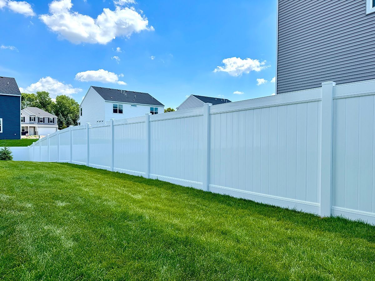 A bright, white vinyl privacy fence runs across a lush green lawn under a sunny, blue sky with houses in the background.