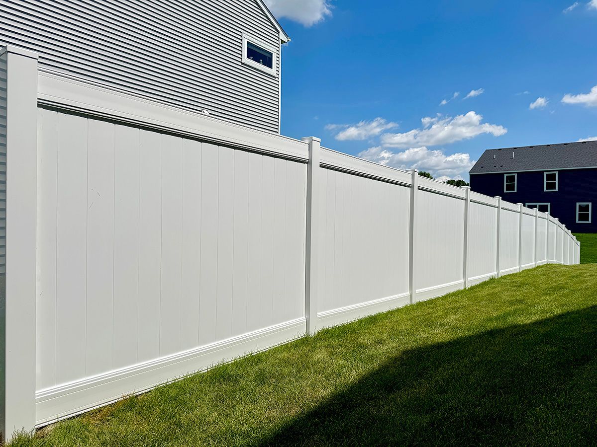A tall, white vinyl privacy fence lines a grassy yard on a sunny day with houses visible in the background.