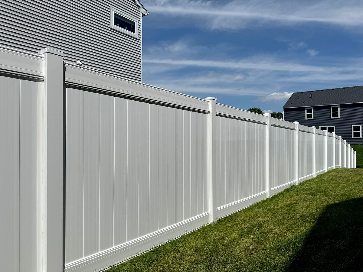 A white vinyl privacy fence runs along a grassy lawn next to residential homes under a blue sky.
