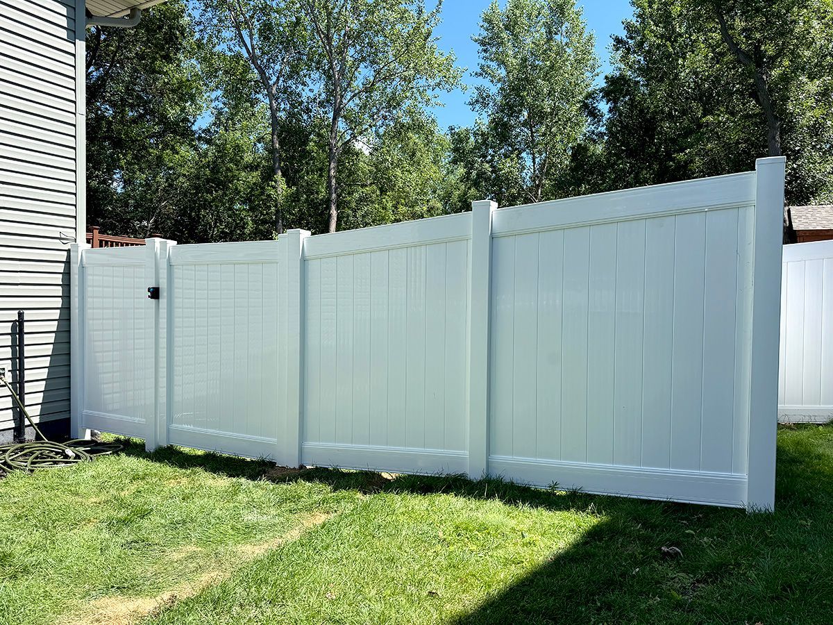 A tall, white vinyl privacy fence stands on a green lawn next to the side of a residential house.