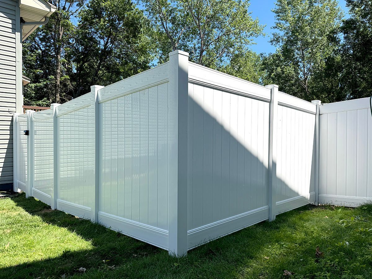 A white vinyl privacy fence with vertical panels and square posts stands on a green lawn against a backdrop of trees.