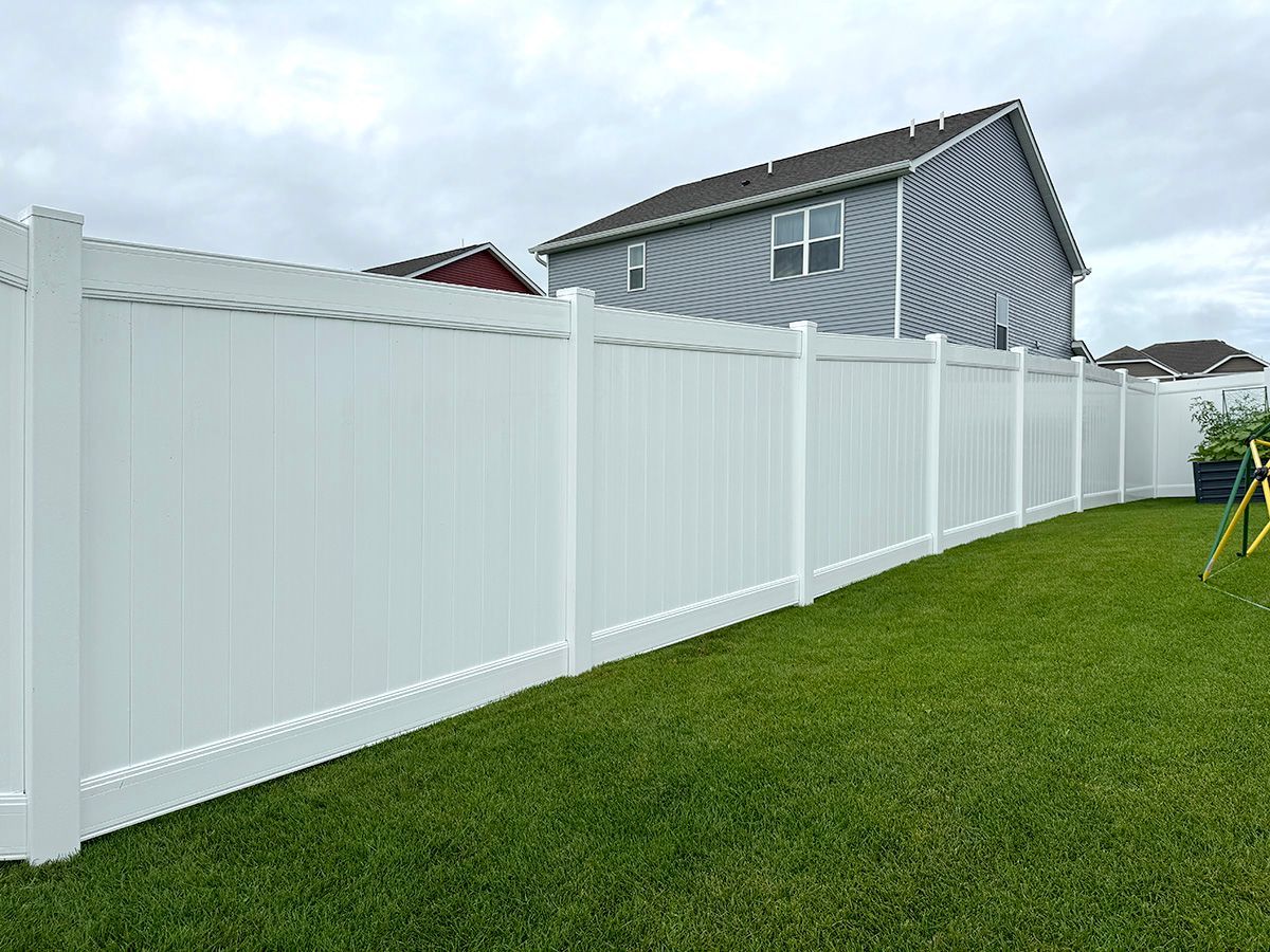 A white vinyl privacy fence stands in a grassy backyard next to a residential house.