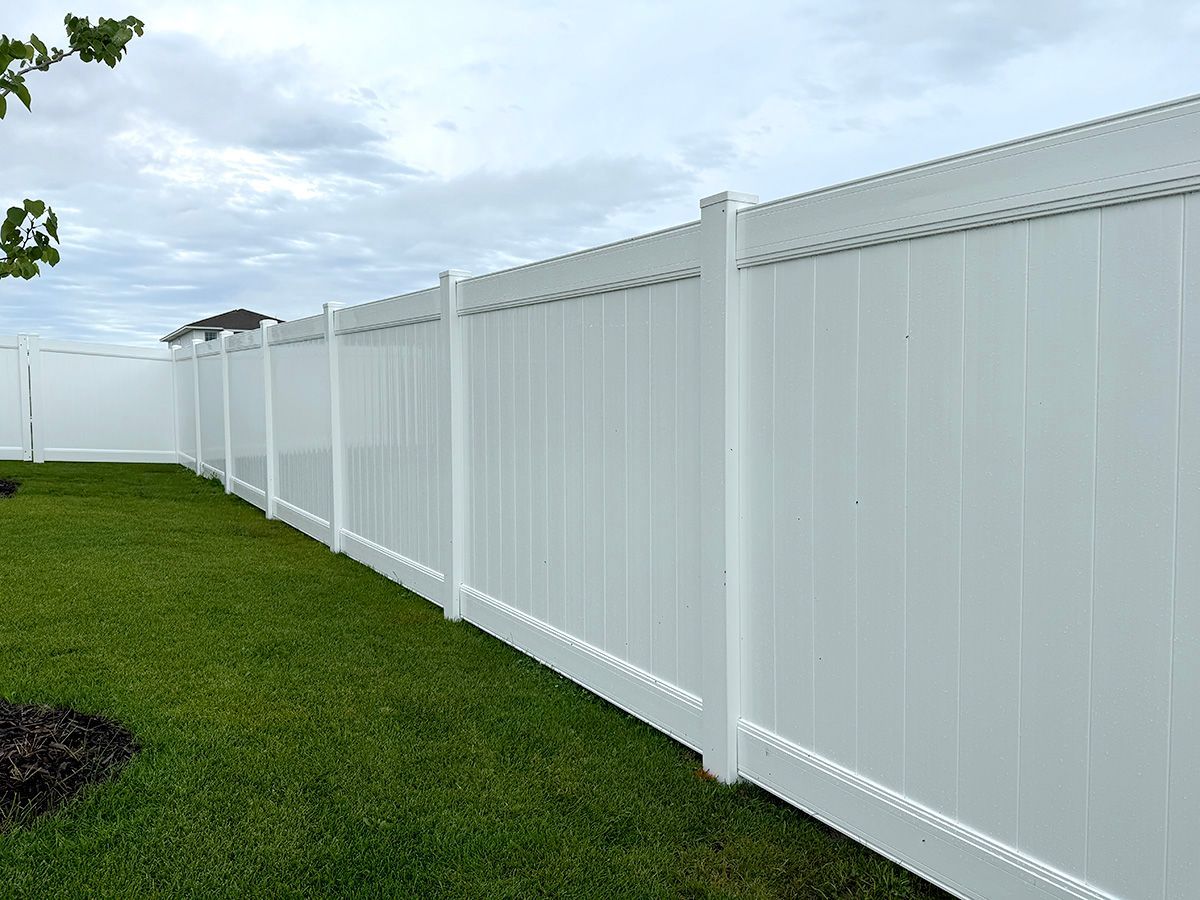 A white vinyl privacy fence lines a grassy backyard under a cloudy sky.