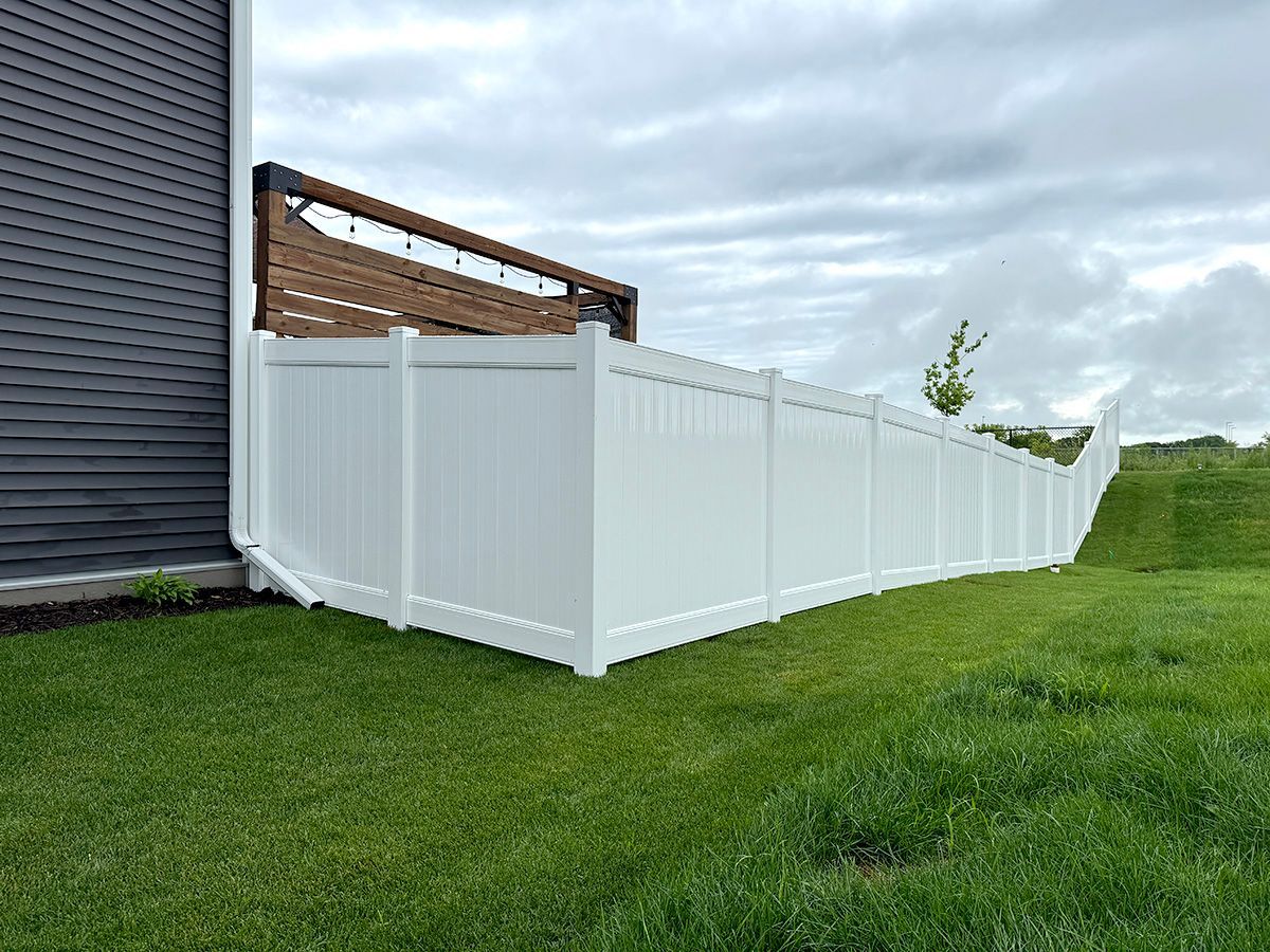 A tall white vinyl privacy fence stands next to a dark gray house wall on a green lawn under a cloudy sky.