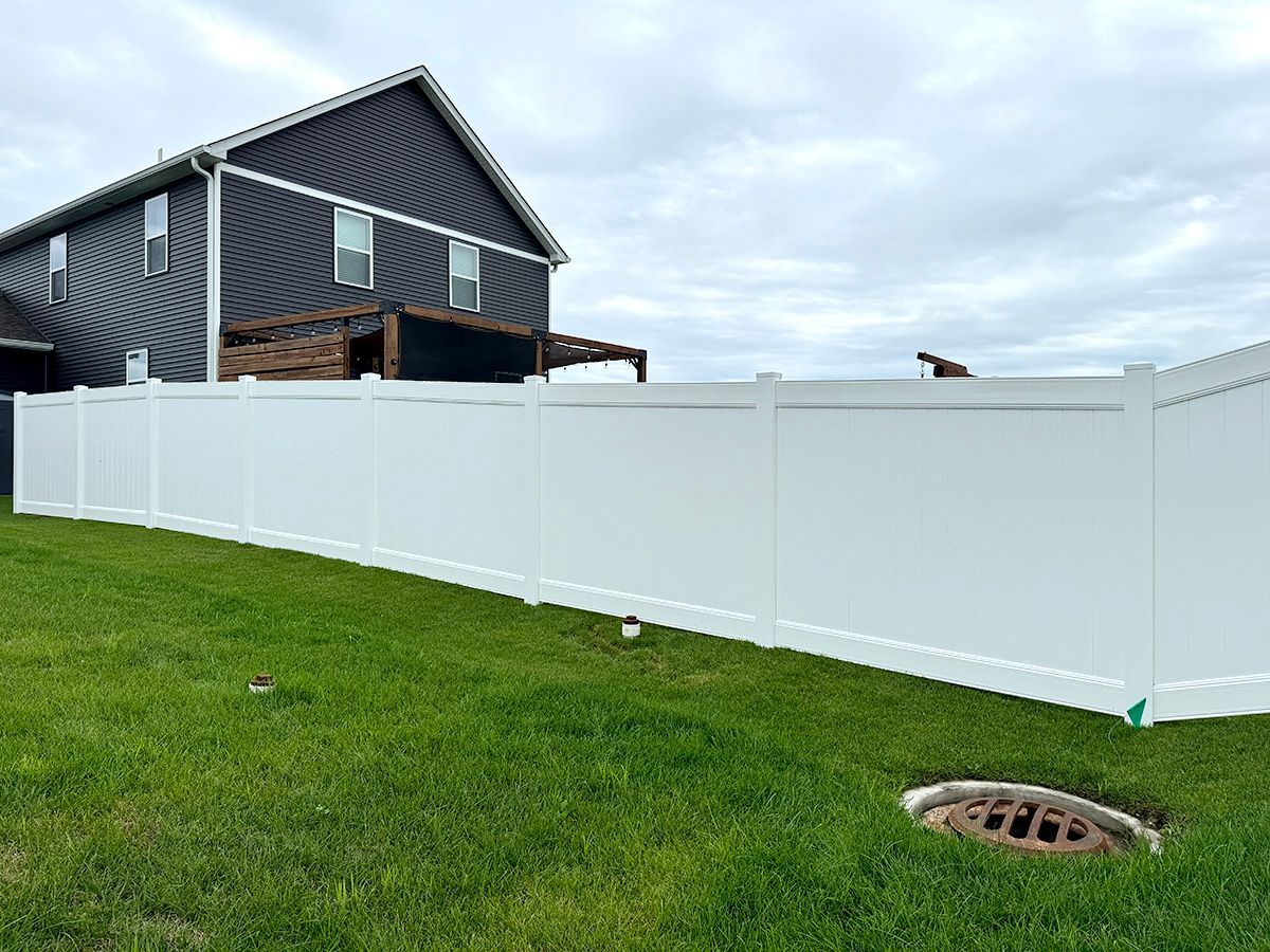 A long white vinyl privacy fence stands in a grassy backyard next to a two-story house with dark siding.