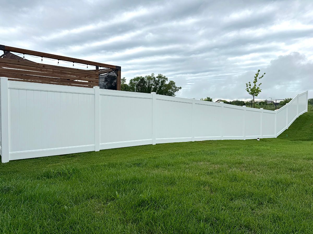 A white vinyl privacy fence runs across a grassy backyard under a cloudy sky.