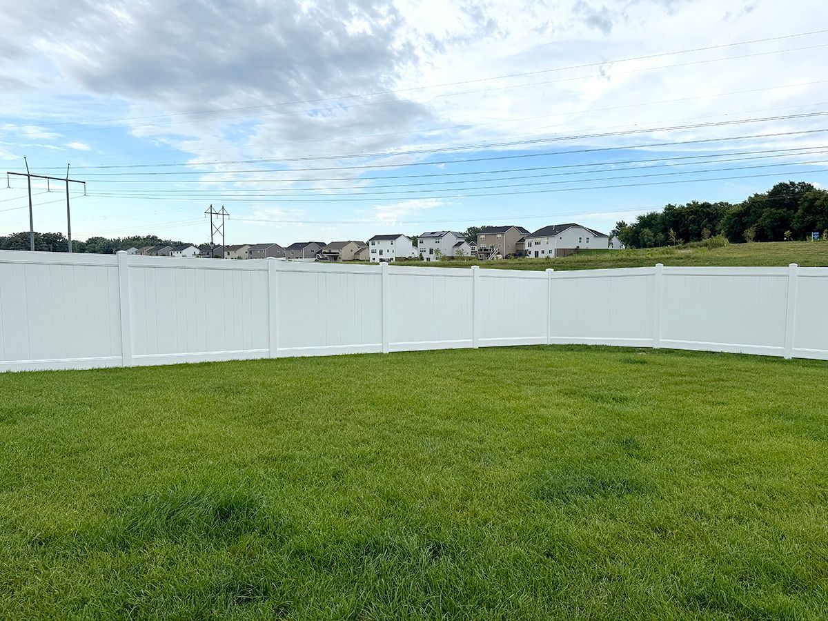 A white vinyl privacy fence encloses a green grassy lawn under a partly cloudy sky, with houses in the distance.
