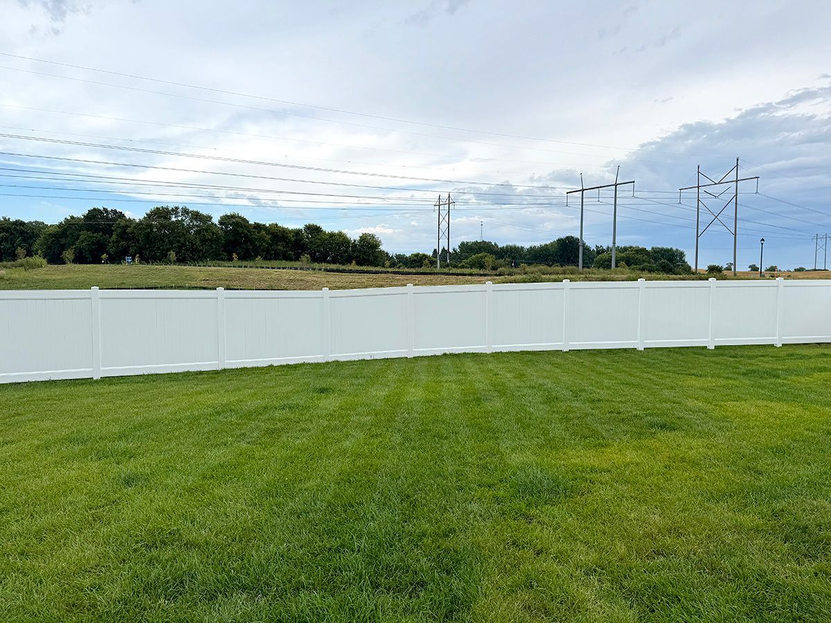 A backyard lawn with a white vinyl privacy fence, set against a backdrop of trees, power lines, and a cloudy sky.