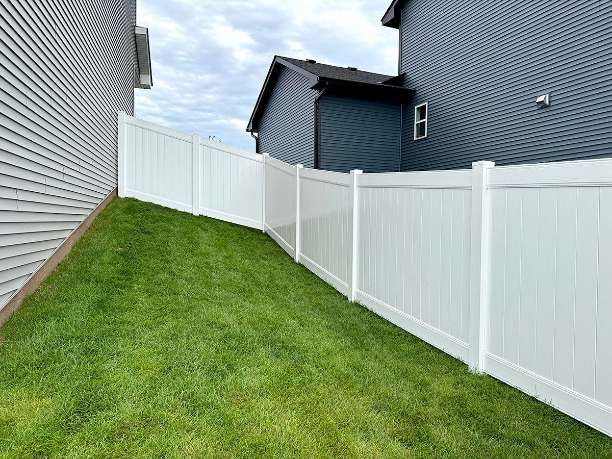 A white vinyl privacy fence runs along a grassy, sloped residential yard between two suburban houses.