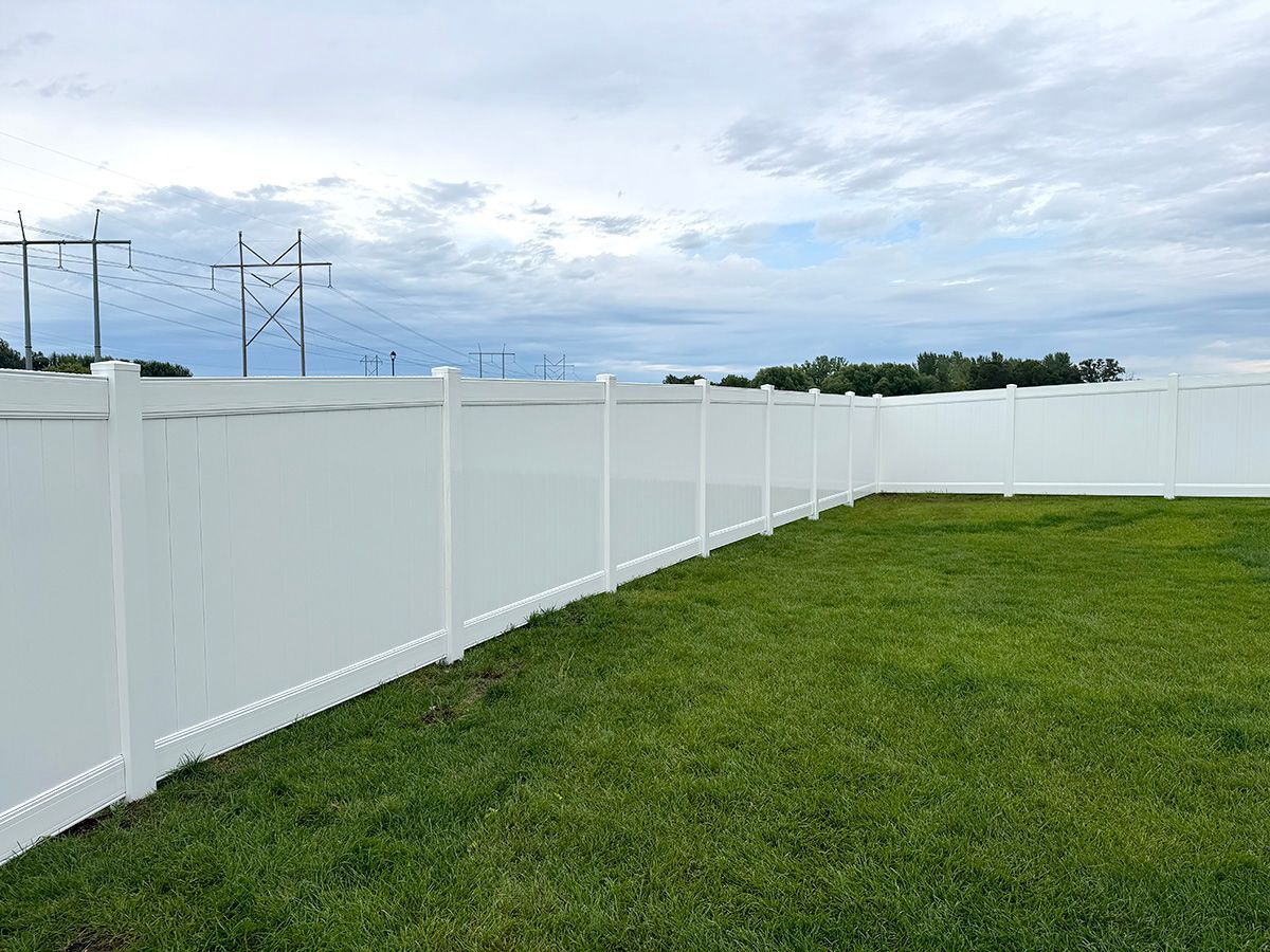 A white vinyl privacy fence encloses a green grassy lawn under a cloudy sky with utility towers in the background.