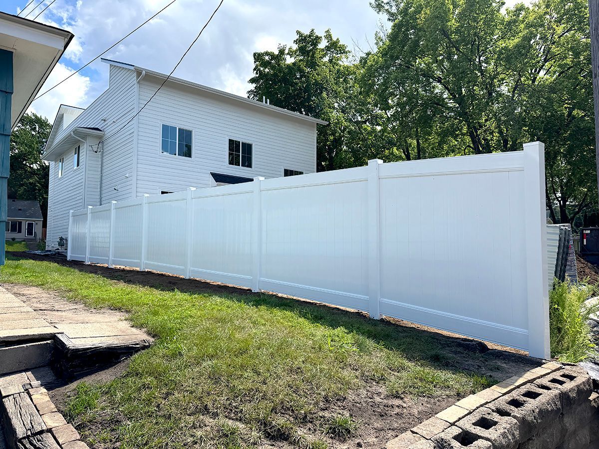 A long, white vinyl privacy fence stands in a grassy yard beside a two-story white house on a sunny day.