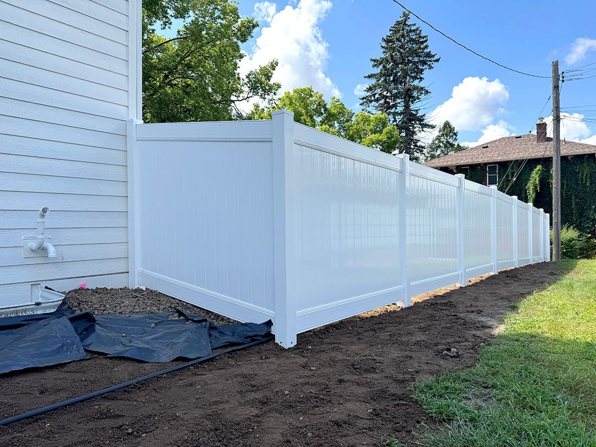 A new white vinyl privacy fence installed next to the side of a house with disturbed soil in the foreground.