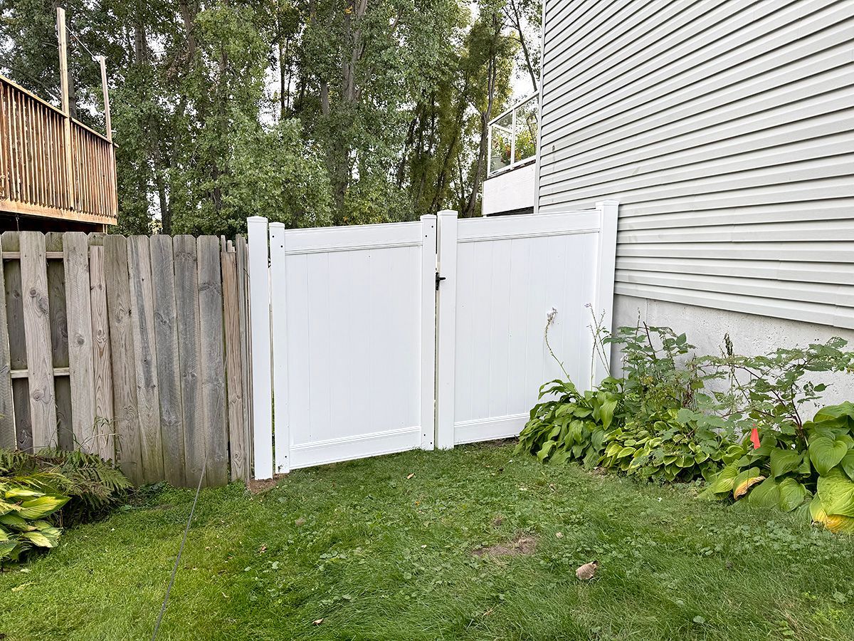 A white vinyl double gate installed in a grassy backyard between a wood fence and a house siding.