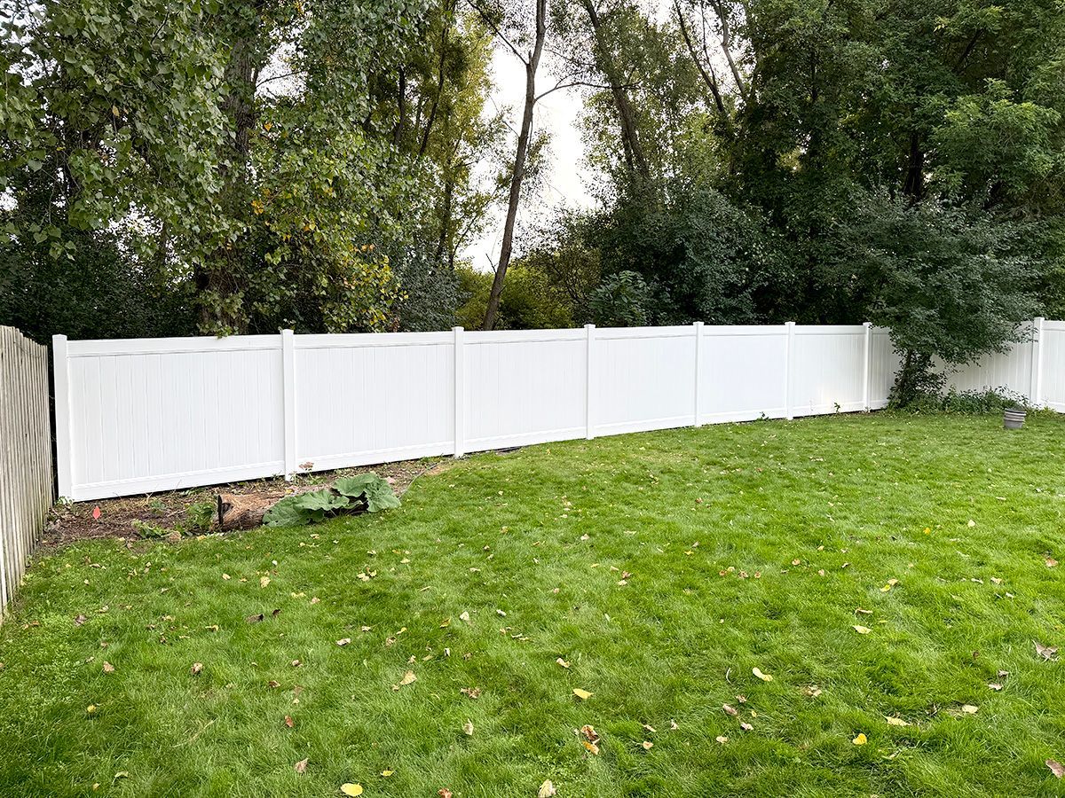 A white privacy fence separates a grassy backyard from a dense line of trees in the background.