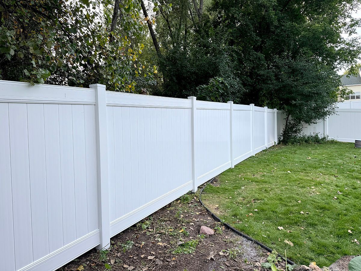 A white vinyl privacy fence stands in a backyard next to a grassy lawn and trees.