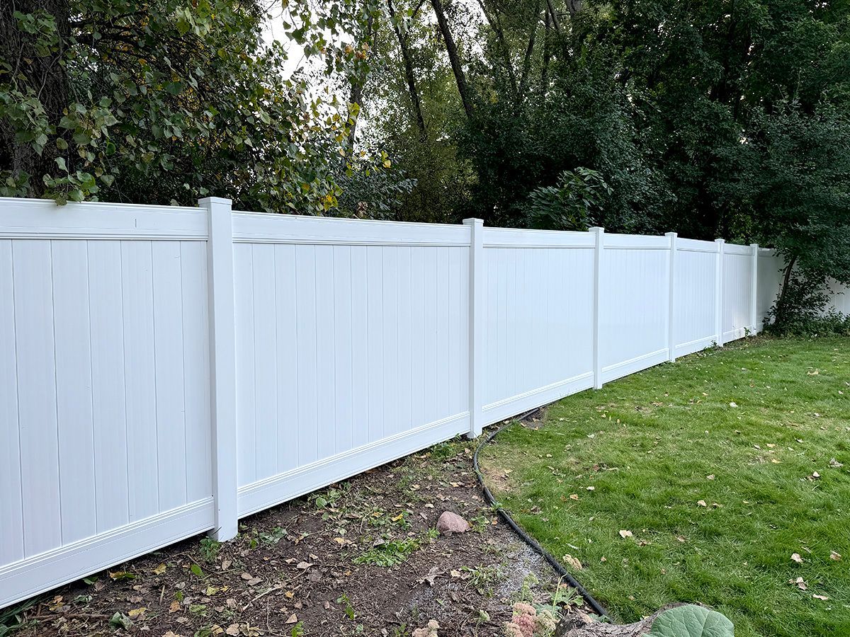 A white vinyl privacy fence lines a grassy yard next to a wooded area.