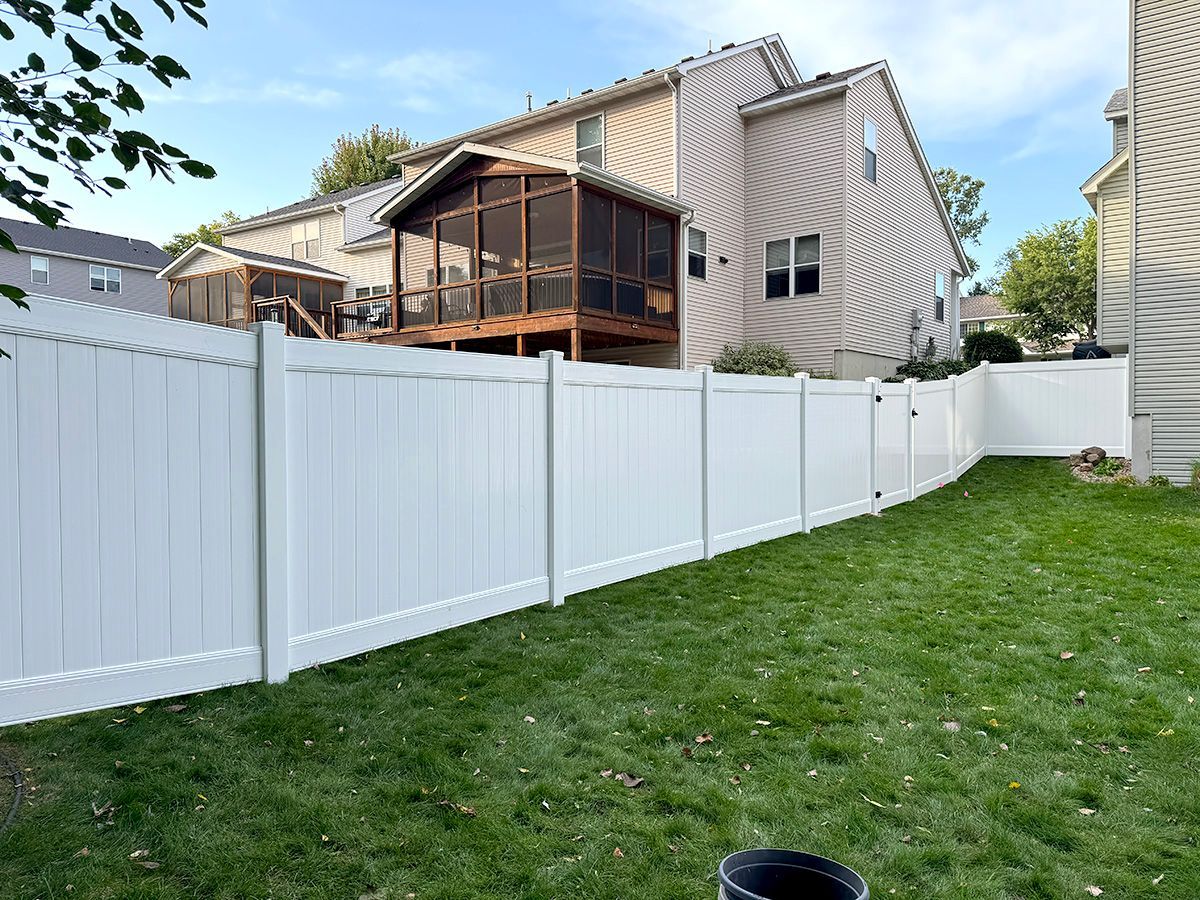 A white vinyl privacy fence curves through a residential backyard, separating the lawn from a beige house with a porch.