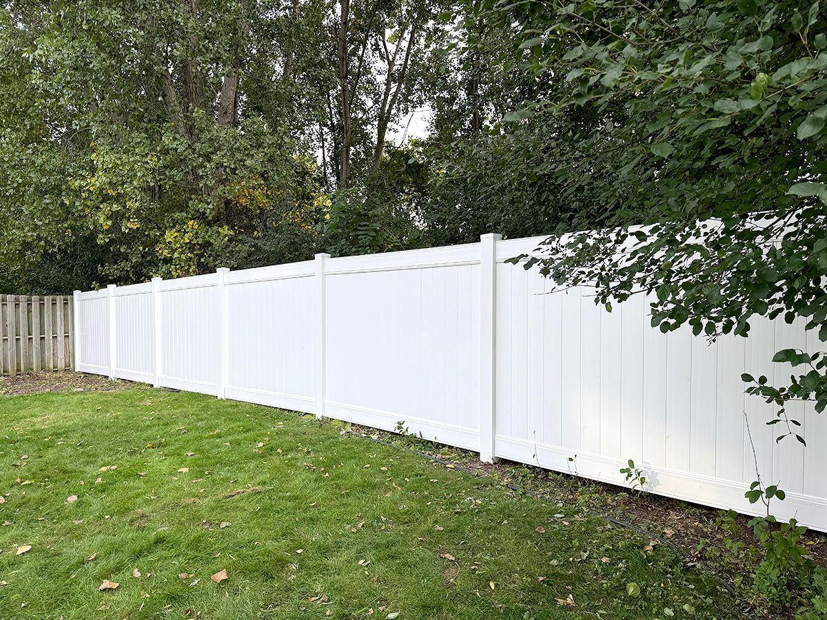 A long, white privacy fence stretches across a green lawn in front of a backdrop of dense green trees.