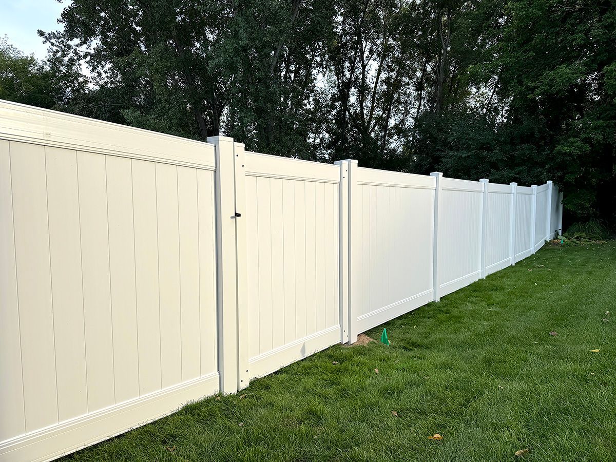 A long, white vinyl privacy fence stands in a grassy yard in front of green trees.