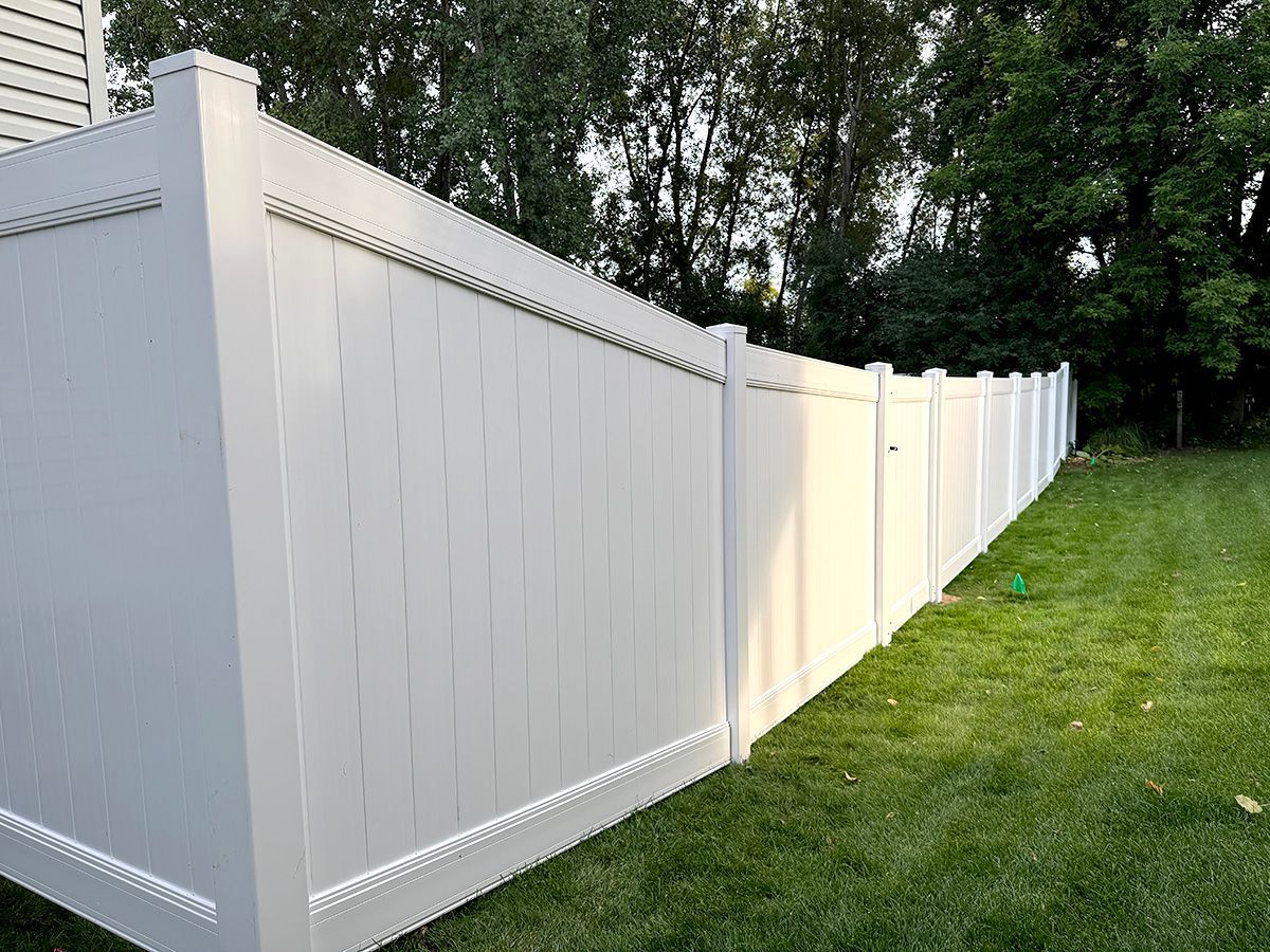 A tall, white vinyl privacy fence stands on a green lawn bordering trees at dusk.