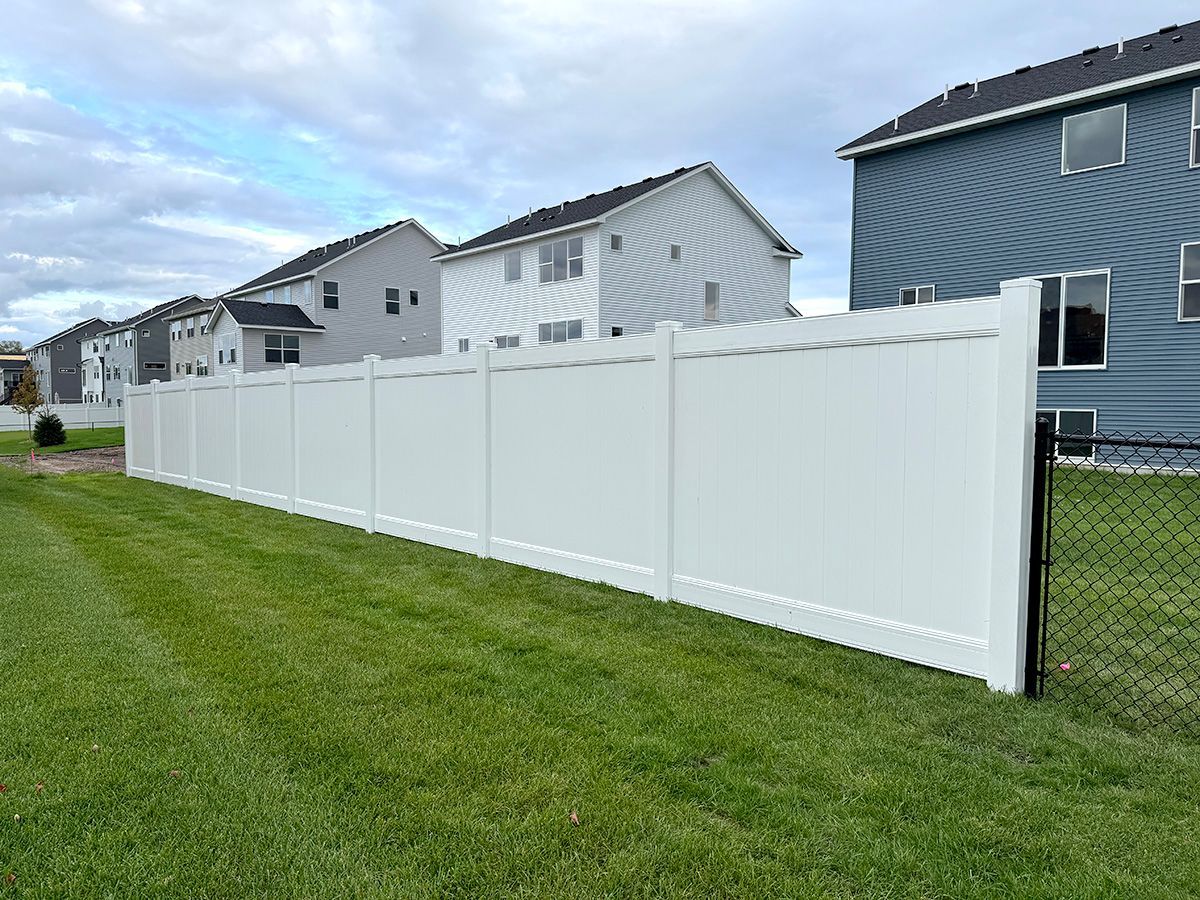 A long white vinyl privacy fence stands in a grassy yard, transitioning to a chain-link section near suburban houses.