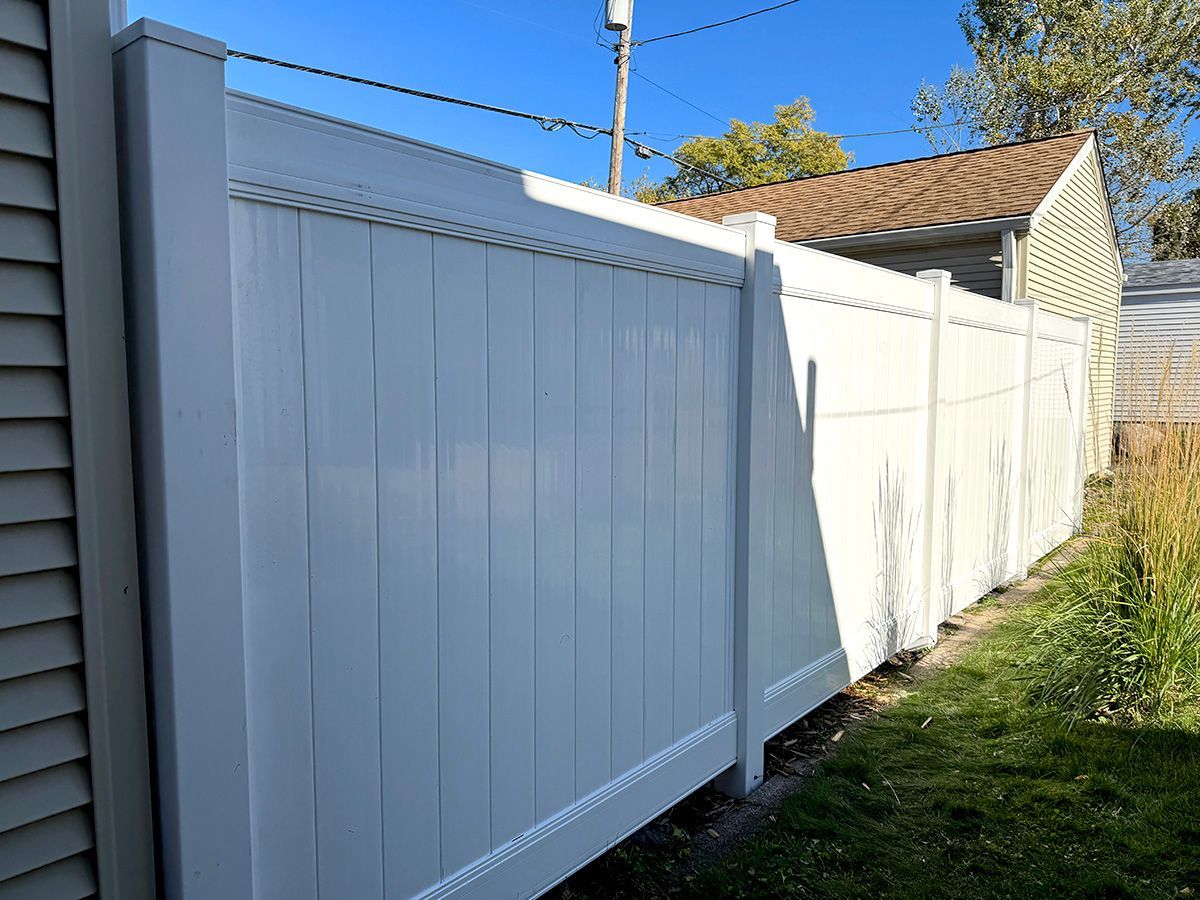 A tall, white vinyl privacy fence stands in a grassy yard next to the side of a house on a sunny day.