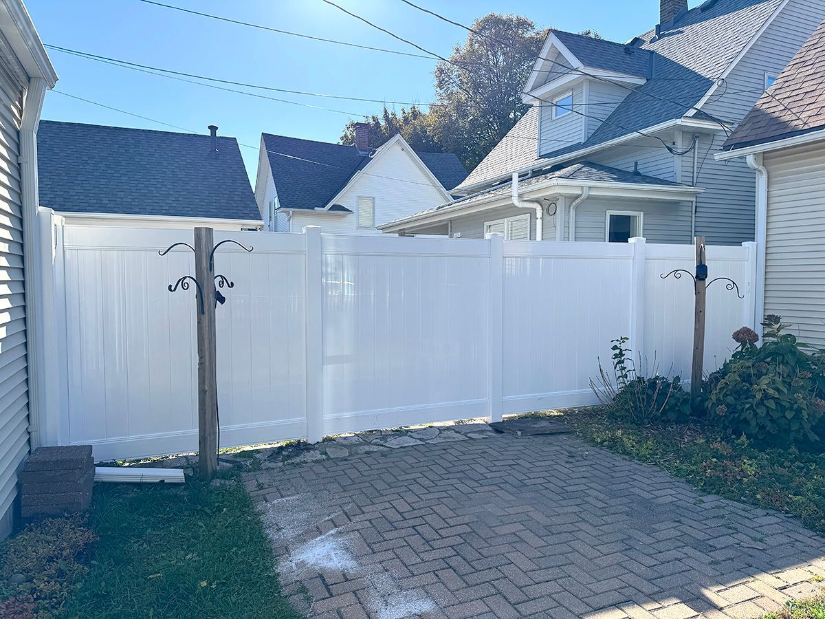 A white vinyl privacy fence separates a brick patio from neighboring houses on a sunny day.