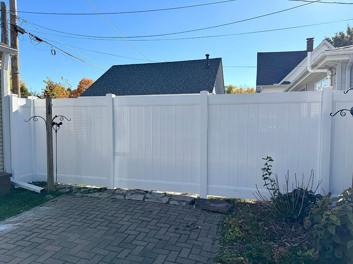 A white vinyl privacy fence stands in a backyard with a brick patio, against a background of houses and blue sky.