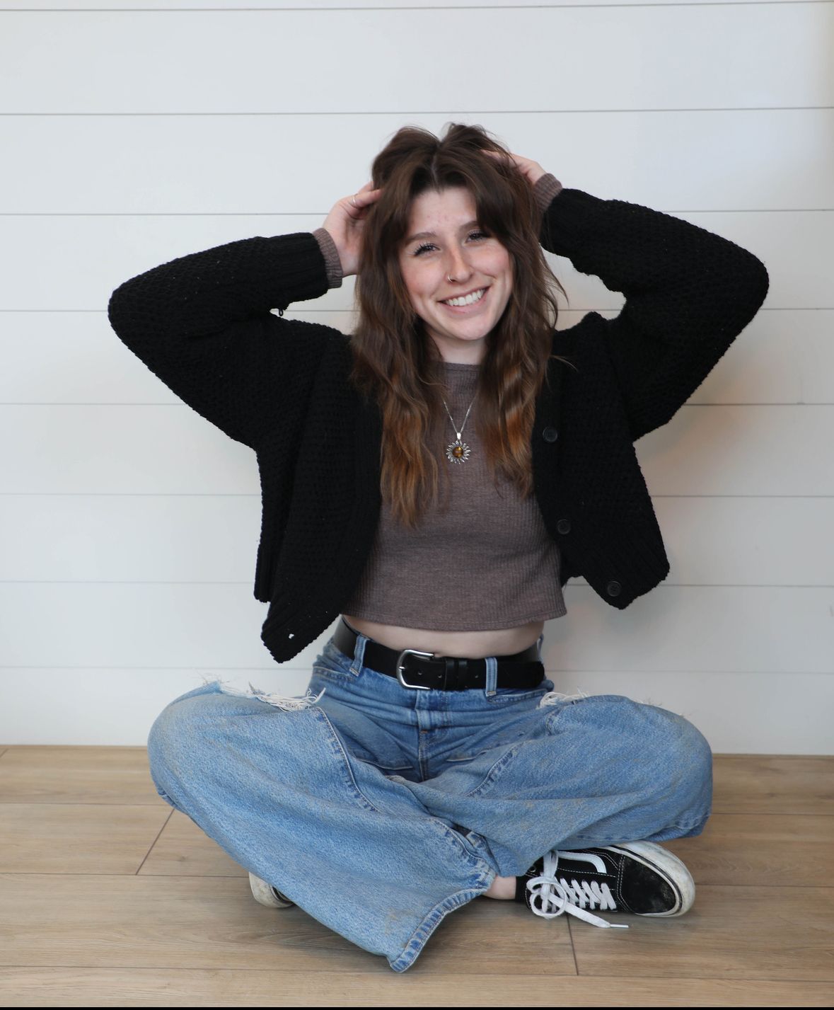 Woman sitting cross-legged, smiling, with hands in hair, wearing a black jacket, brown top, and jeans.