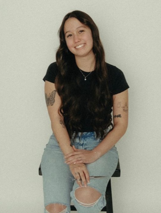 Young woman with long brown hair, smiling, seated, wearing black shirt and ripped jeans, tattoos on arms.