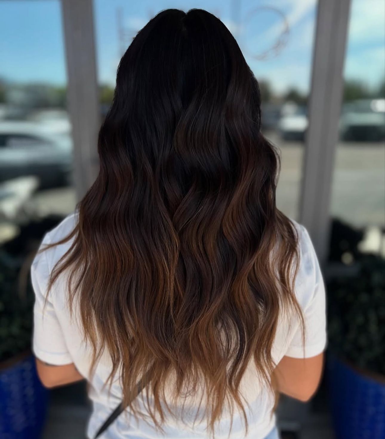 Woman with long, wavy hair in a brown ombre style, standing outdoors in front of a window.