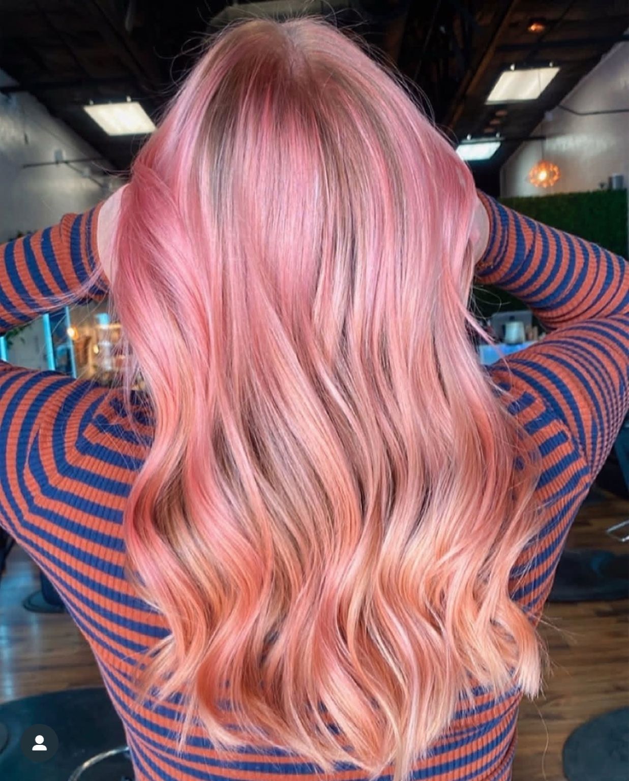 Woman with long wavy pink-to-peach ombre hair, holding hands up. Back view, indoors.
