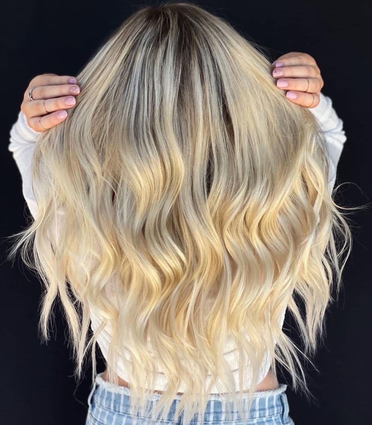 Woman with long, wavy blonde hair, holding it out with both hands, against a dark background.
