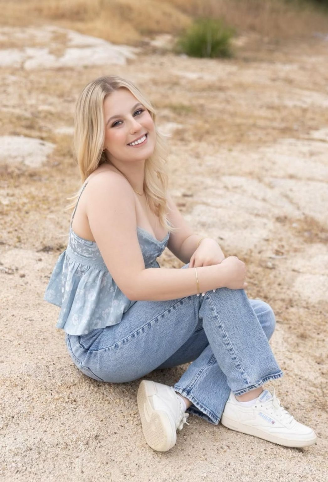 Blonde woman in blue top and jeans, smiling while sitting on the ground.