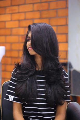 Young woman with long, layered, black hair smiling, wearing striped shirt, in front of brick wall.